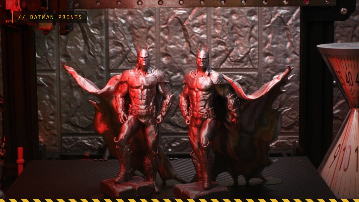 Two 3D printed Batman figures with capes on a printer bed under red light, by iczfirz
