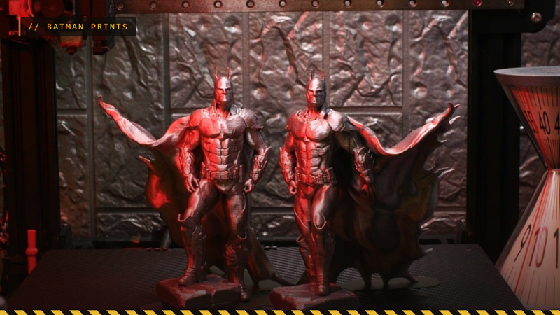 Two 3D printed Batman figures with capes on a printer bed under red light, by iczfirz